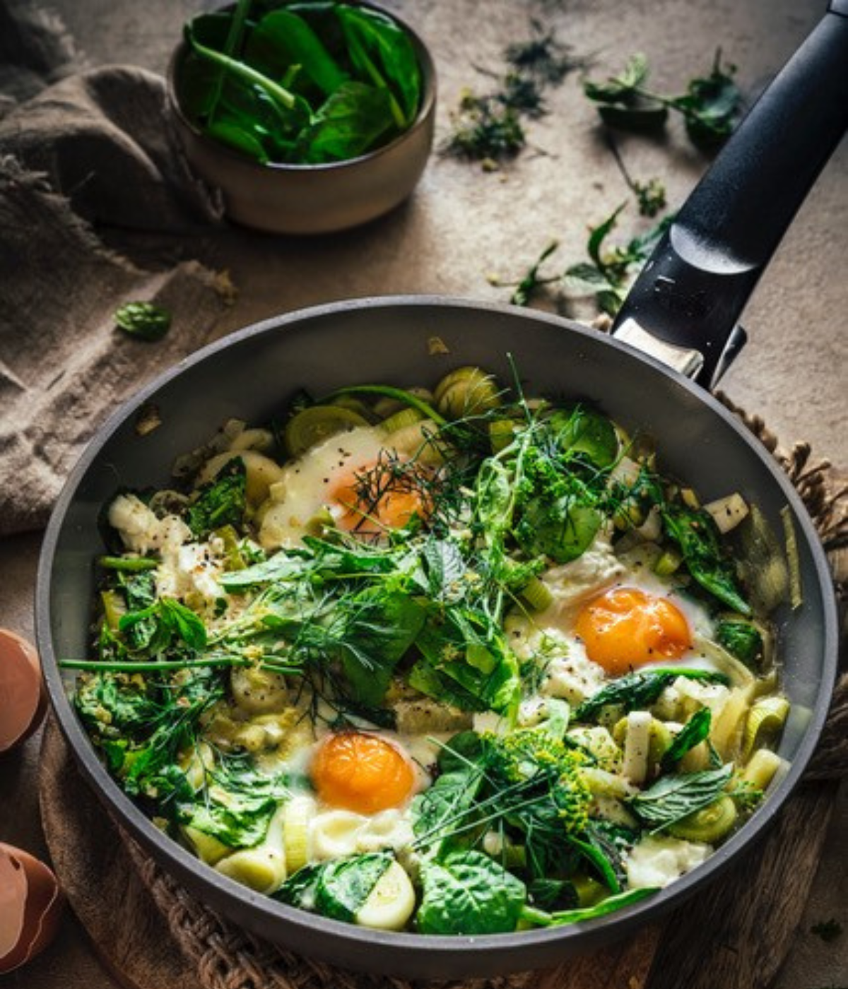 Eine Fissler-Pfanne mit sautiertem Grünzeug, Kräutern und drei Spiegeleiern. Frischer Spinat ziert die Platte; abgebildetes Kochgeschirr.