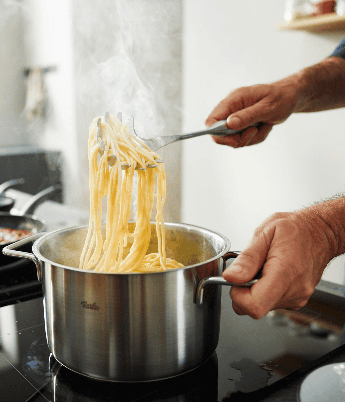 Ein Hobbykoch hebt Spaghetti mit einer Zange aus einem Fissler-Edelstahltopf, der deutsche Präzision und zeitloses Design demonstriert.