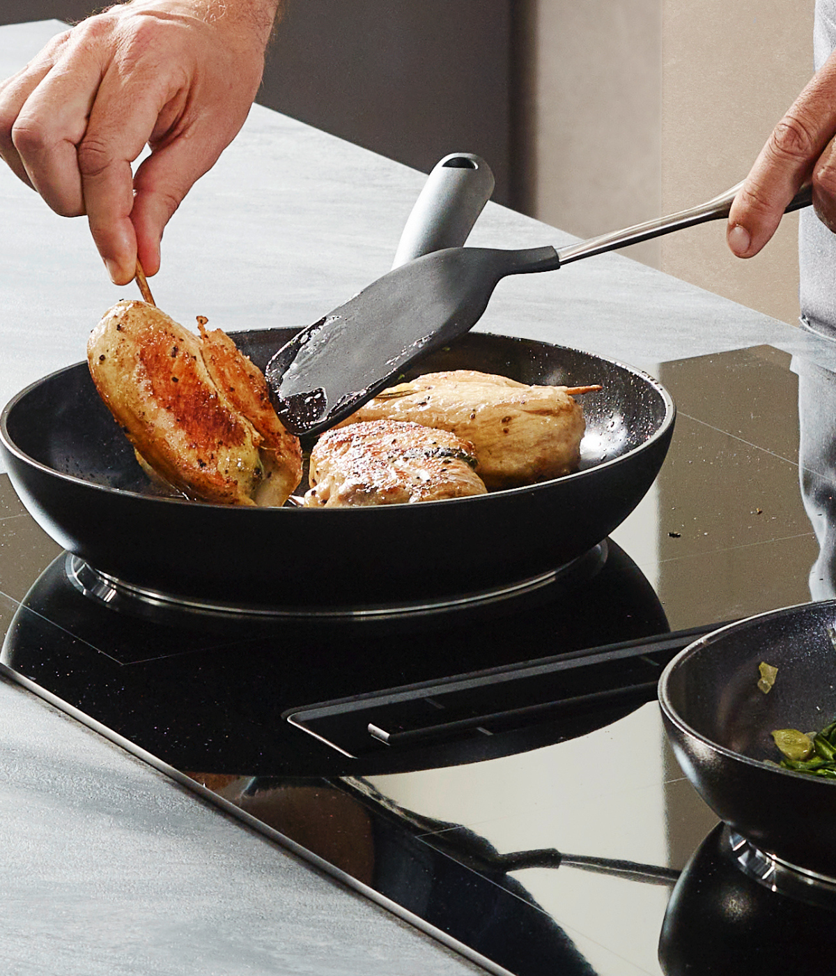 Ein Koch bereitet ein gefülltes Hähnchen in einer eleganten Fissler-Pfanne auf einem Elektroherd zu; daneben steht eine Fissler-Pfanne mit frischem Grün.