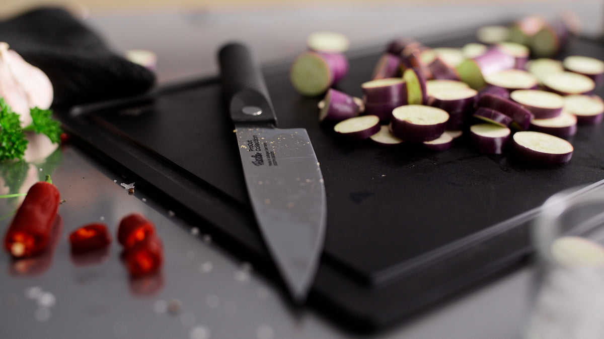 Fissler ProCut Kochmesser 16cm auf schwarzem Brett mit geschnittener Aubergine, Chili, Knoblauch & Petersilie - Präzision trifft auf modernes deutsches Design.