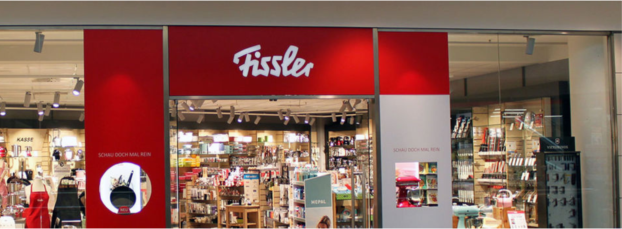 Das Fissler-Geschäft mit der charakteristischen rot-weißen Fassade präsentiert hochwertiges, in Deutschland hergestelltes Edelstahl-Kochgeschirr in einem hellen Einkaufszentrum.