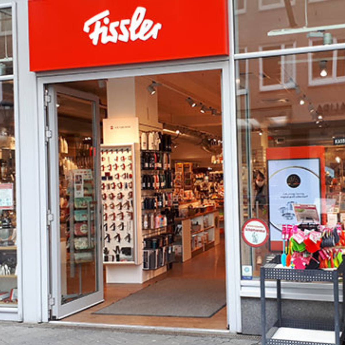 Fissler-Schaufenster mit dem ikonischen roten Schild, offenen Glastüren und hochwertigem, in Deutschland hergestelltem Kochgeschirr, das innen und außen präsentiert wird.
