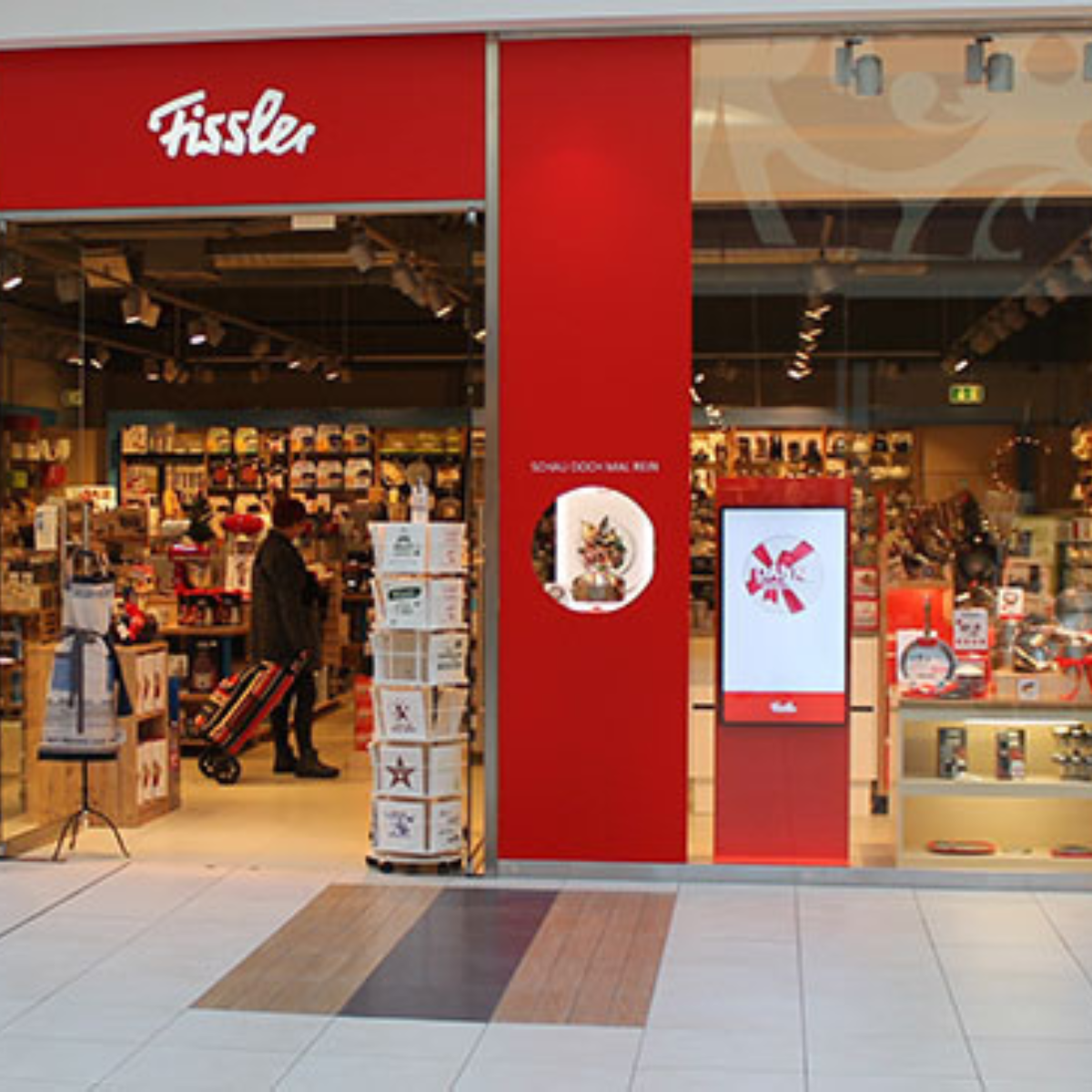 Fissler-Geschäft mit dem charakteristischen roten Eingang, Glastüren und hochwertigem deutschem Kochgeschirr in der Auslage. Shopper mit Einkaufswagen vor dem Geschäft.