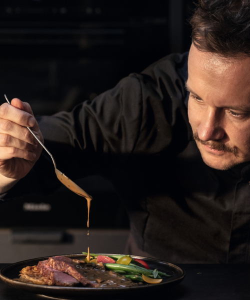 Ein Koch verwendet Fissler-Kochgeschirr, um ein Gourmetgericht auf einem dunklen Teller kunstvoll mit Sauce zu beträufeln und so Präzision und Qualität zu demonstrieren.