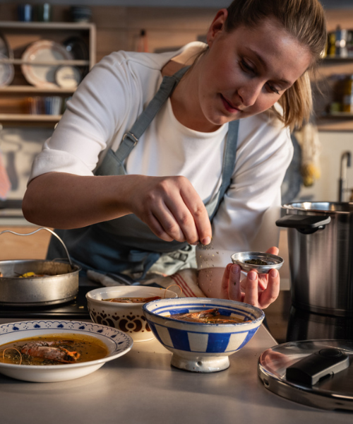 Eine Frau in einer Schürze kocht in ihrer Küche eine Suppe mit hochwertigen Töpfen und Kochgeschirr aus rostfreiem Stahl von Fissler und lächelt während des Kochens.