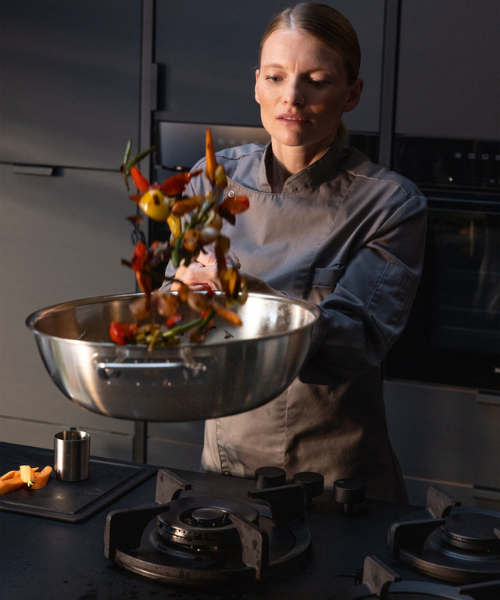 Ein Koch in grauer Uniform schwenkt fachmännisch Gemüse in einer Fissler-Edelstahlpfanne auf einem schlichten, modernen Herd.