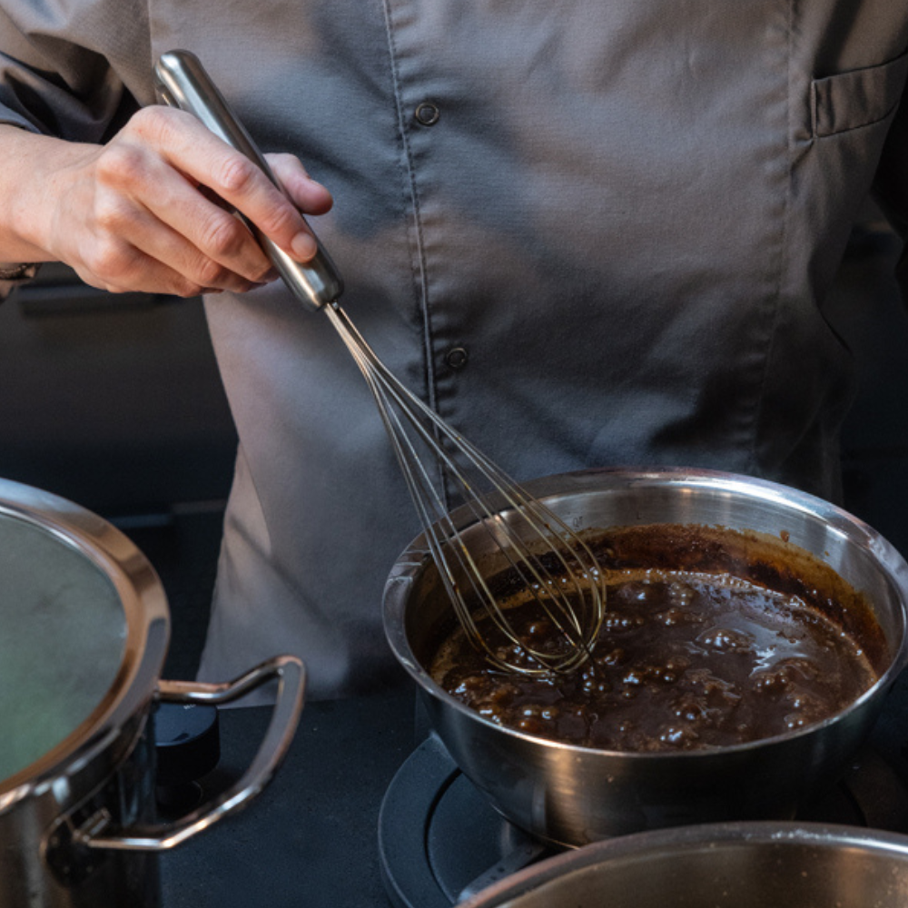 Ein Koch in Grau verwendet einen Fissler-Topf, um eine Soße auf dem Herd zu verquirlen; ein weiterer hochwertiger Fissler-Topf mit Deckel steht daneben.