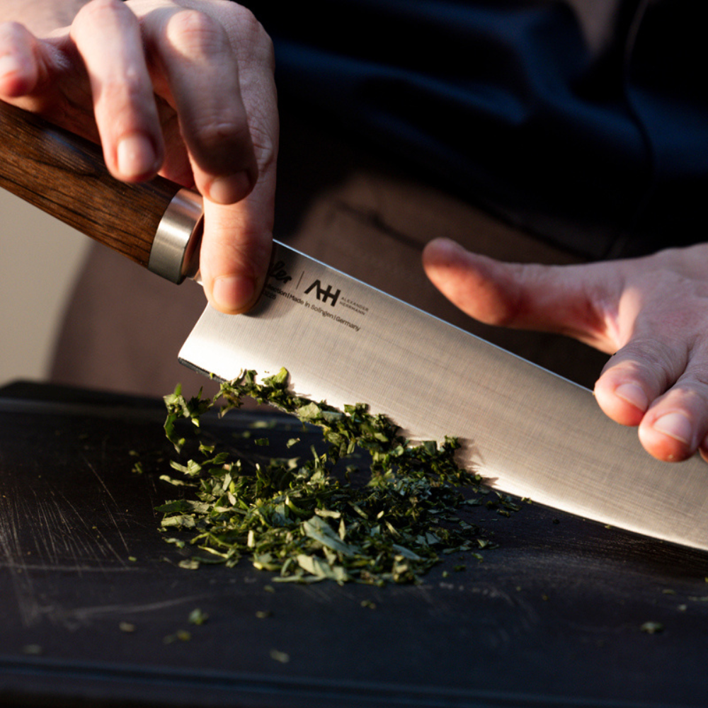 Eine Person hackt frische Kräuter auf einem schwarzen Schneidebrett mit einem Fissler-Kochmesser mit ergonomischem Holzgriff fein.