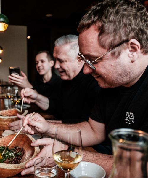 Drei Männer genießen gemeinsam eine Mahlzeit an einem Restauranttisch; jeder von ihnen lächelt, während er Essstäbchen benutzt. Ein Mann hält den Moment mit seinem Handy fest. Auf dem Tisch stehen Gläser mit Wein und Wasser neben meisterhaft zubereiteten Gerichten, die Fisslers Engagement für Präzision und zeitloses Design widerspiegeln. Die Szene unterstreicht das gesellige Beisammensein beim Essen – ermöglicht durch hochwertiges Kochgeschirr aus der renommierten Heritage-Kollektion von Fissler.