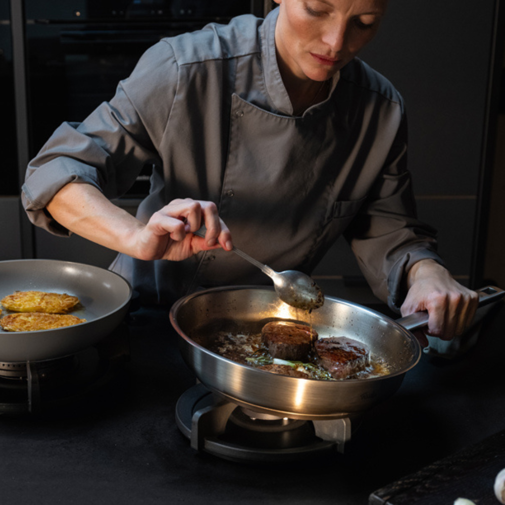 Ein Koch brät Fleisch in einer Fissler-Edelstahlpfanne an und löffelt Sauce, während eine andere hochwertige Fissler-Pfanne das Essen auf einem modernen Herd kocht.