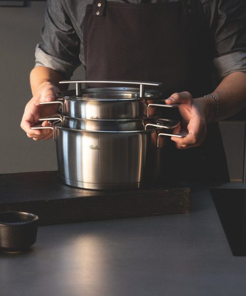 Eine Person stapelt Fissler-Edelstahltöpfe mit Deckeln auf einem Küchentisch, um den Glanz und das zeitlose Design aus deutscher Produktion zu präsentieren.