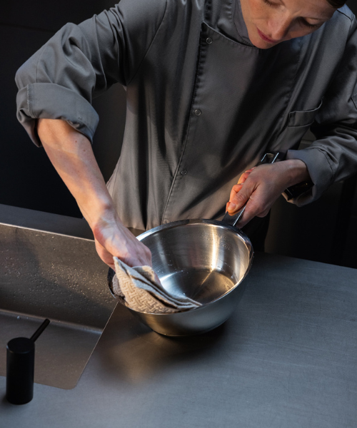 Ein Koch in einer grauen Jacke trocknet eine Fissler-Edelstahlschüssel mit einem gemusterten Tuch an einer glatten Metalltheke in der Nähe der Spüle ab.