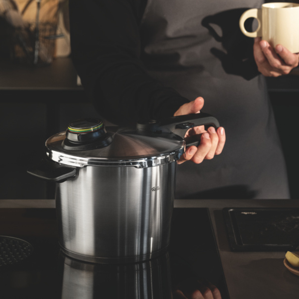 Eine Person steht hinter einem Fissler Schnellkochtopf aus Edelstahl auf einem Küchentisch, der deutsche Qualität und zeitloses Design präsentiert.