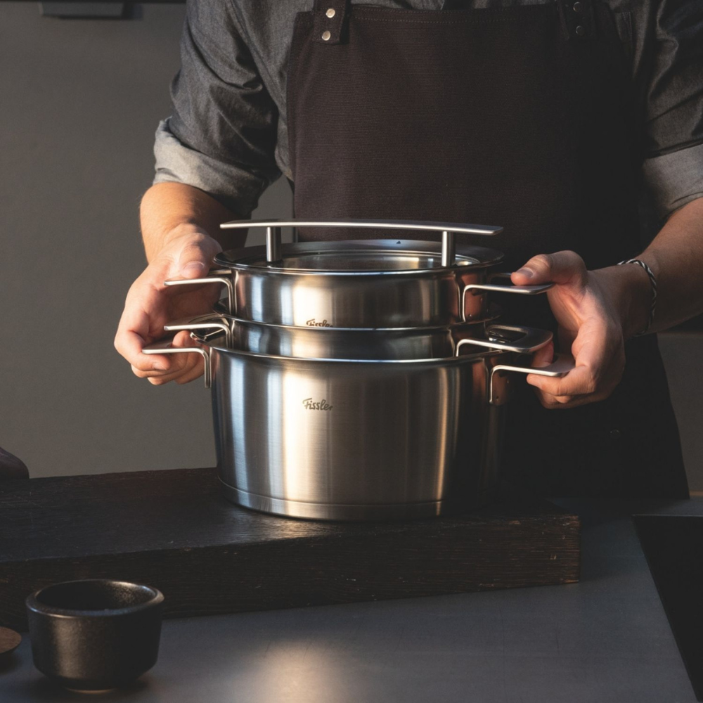 Eine Person in dunkler Schürze stapelt Fissler-Edelstahltöpfe mit Deckeln auf einer hölzernen Unterlage, die deutsche Präzision und Design demonstriert.