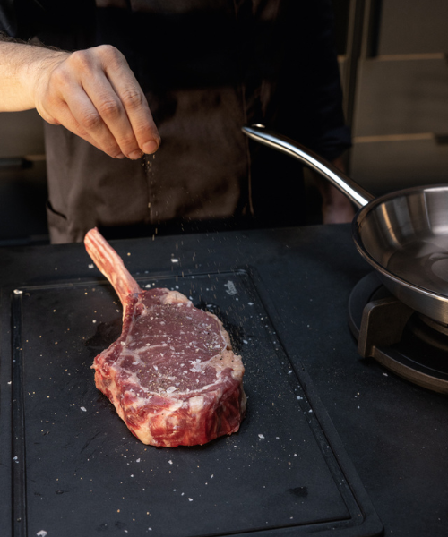 Eine Person würzt ein Tomahawk-Steak auf einem Schneidebrett neben einer Fissler-Bratpfanne, die das deutsche Präzisionskochgeschirr hervorhebt.