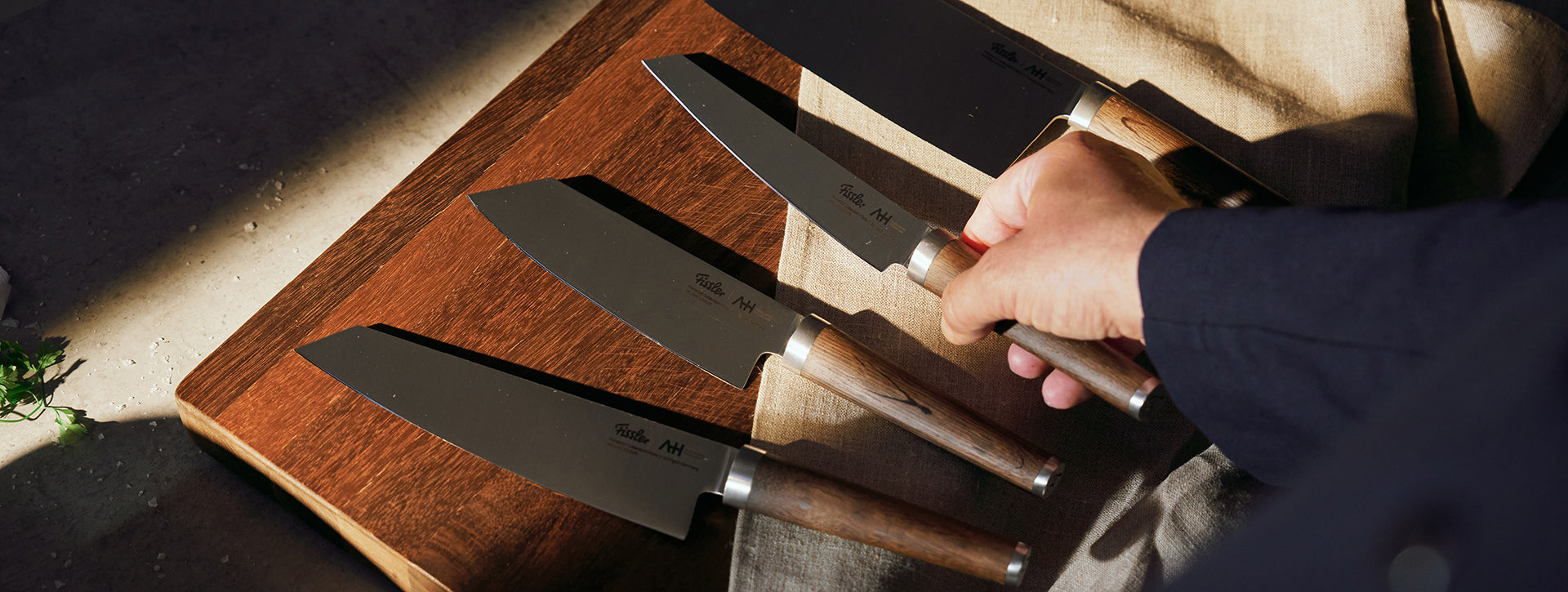 Nahaufnahme einer Hand, die ein Fissler-Kochmesser mit Holzgriff aus vier auf einem Holzbrett liegenden Messern auswählt, wobei die Sonne die Handwerkskunst hervorhebt.