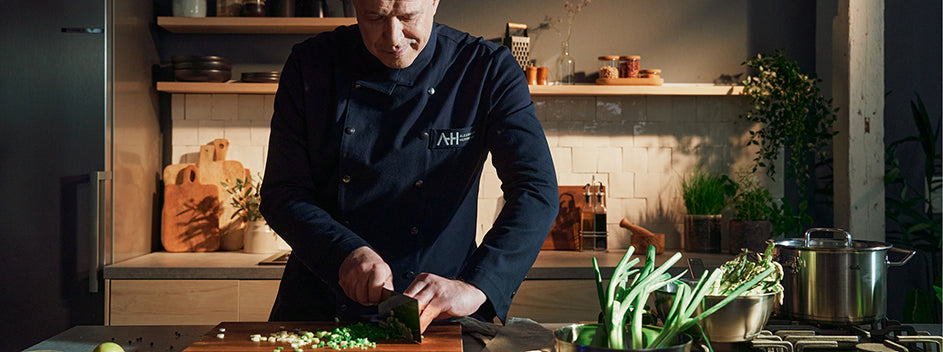 Ein Koch hackt Kräuter auf einem Holzbrett in einer modernen Küche mit einem Fissler-Edelstahltopf, frischem Gemüse und Utensilien im Blick.