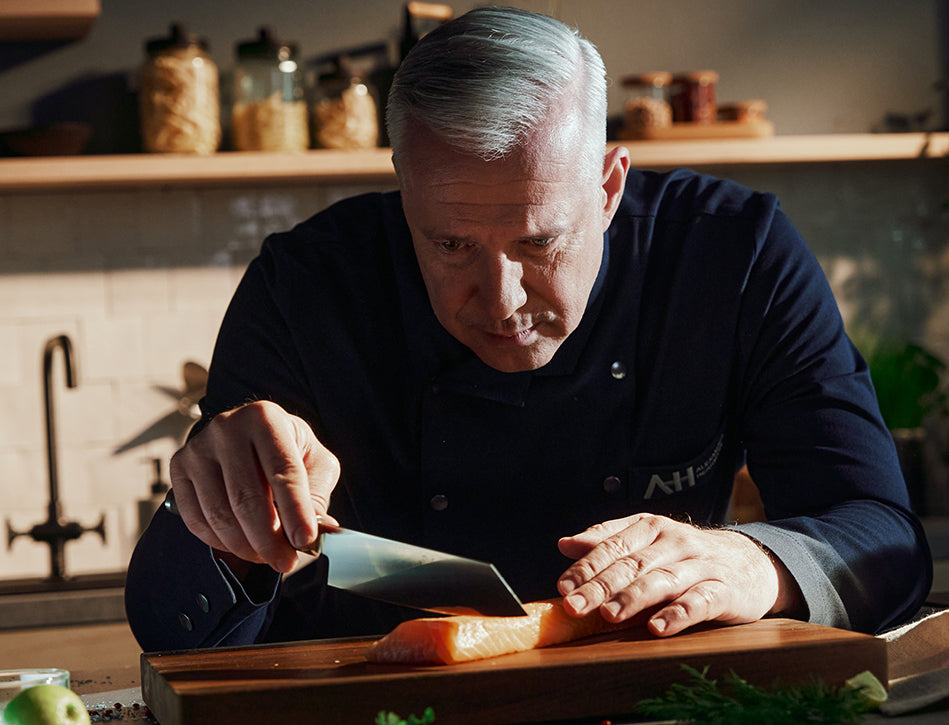Ein Koch mit grauen Haaren schneidet in einer modernen Küche mit hochwertigem Fissler-Kochgeschirr Lachs auf einem Holzbrett in feine Scheiben.
