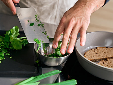 Eine Person verwendet ein präzises Fissler-Kochmesser, um Koriander in eine Edelstahlschüssel zu geben; daneben liegen Brotscheiben.