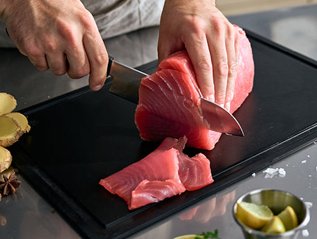 Auf einem Fissler-Schneidebrett schneidet eine Person fachmännisch rohen Thunfisch in Scheiben; Ingwer, Zitronenscheiben und eine Schüssel ergänzen den hochwertigen Arbeitsplatz.
