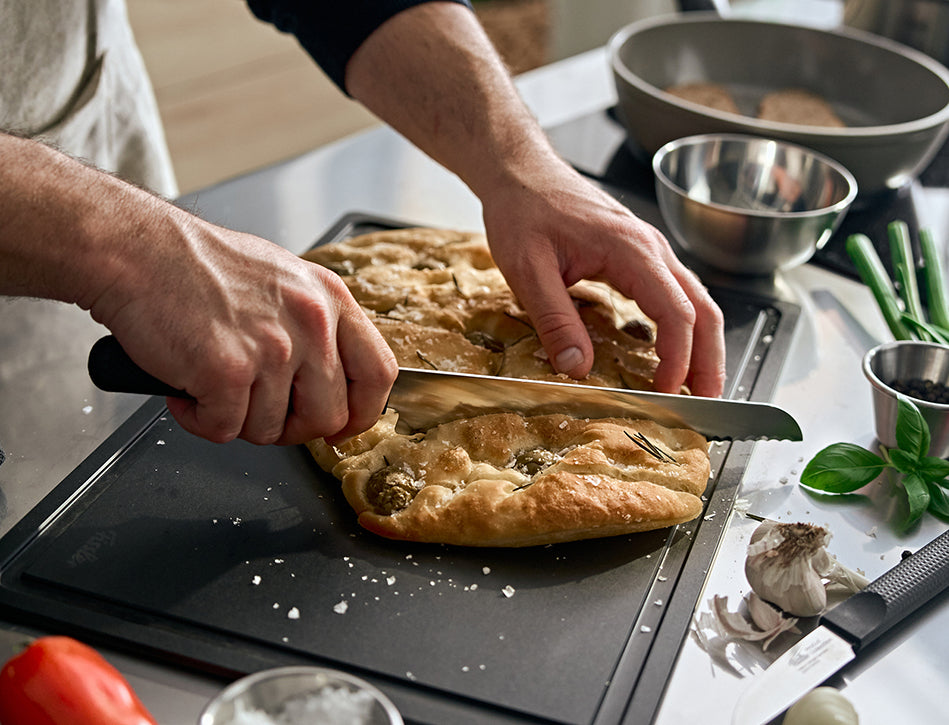Eine Person schneidet Focaccia auf einem Brett, umgeben von Fissler-Edelstahlkochgeschirr, Kräutern und Küchengeräten auf der Arbeitsplatte.
