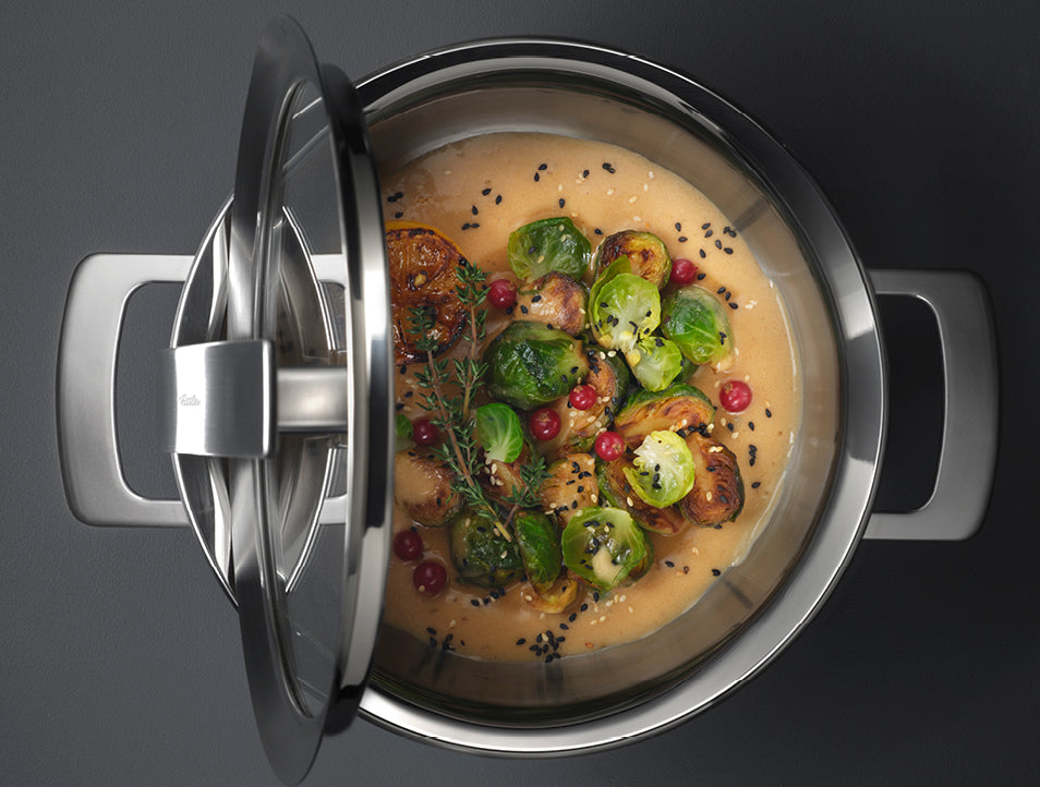 Ein Fissler-Edelstahltopf mit Glasdeckel, gefüllt mit Sahnesauce, Rosenkohl und Preiselbeeren auf einer dunklen Oberfläche.