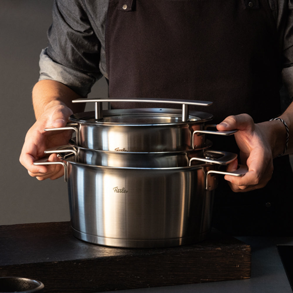 A person in a dark apron stacks two Fissler stainless steel pots with ergonomic handles on a sleek, dark surface, highlighting the brand’s renowned German precision and timeless design.