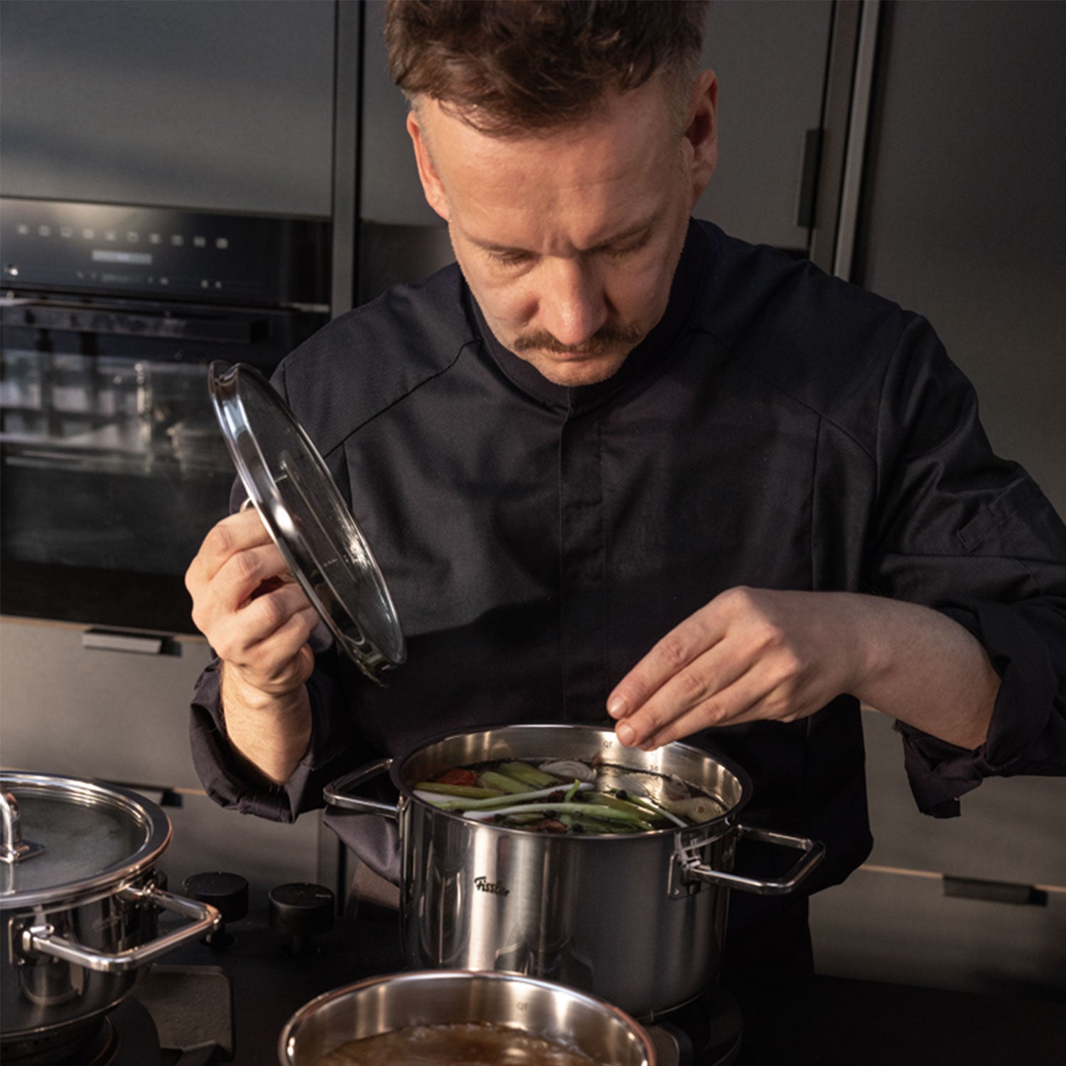 Ein Mann in einer Kochjacke gibt Zutaten in einen Fissler-Edelstahltopf und präsentiert damit deutsche Qualität in einer schlichten, modernen Küche.