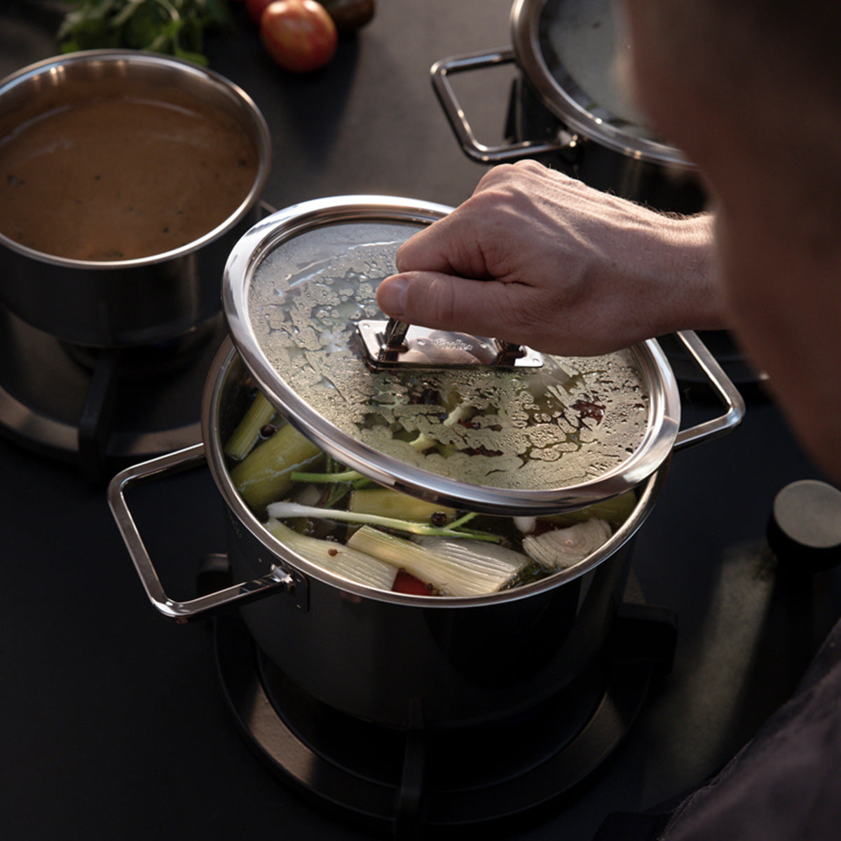 Eine Hand hebt den Fissler Viseo® Pro Glasdeckel 20 cm aus einem Viseo Pro Topf; ein anderer Fissler Topf mit Sauce köchelt neben frischem Gemüse.