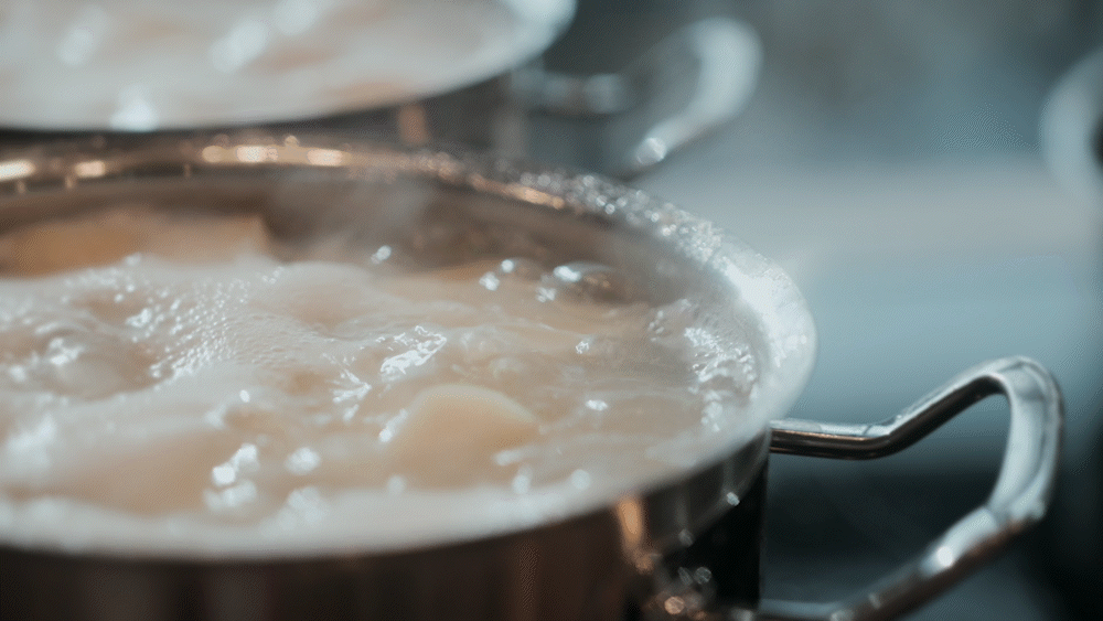 Close-up, high-angle view of two premium Fissler stainless steel pots on a stove, each filled with bubbling water and perfectly sliced potatoes. Steam elegantly rises above the precision-engineered cookware, creating a soft-focus effect that highlights the durability and timeless German design.