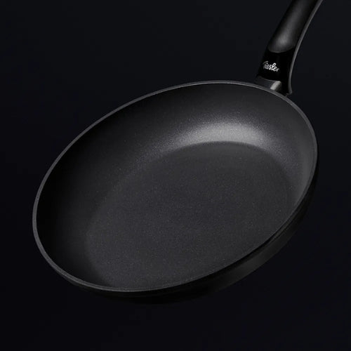 Fissler Antihaft-Bratpfanne mit gebogenem Griff, hergestellt in Deutschland, abgebildet auf schwarzem Hintergrund. Die glatte, leere Innenseite unterstreicht die Qualität.