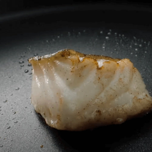 Ein weißes Fischfilet wird in einer Fissler-Pfanne goldbraun gebraten, wobei die Öltropfen die Präzision deutscher Handwerkskunst unterstreichen.