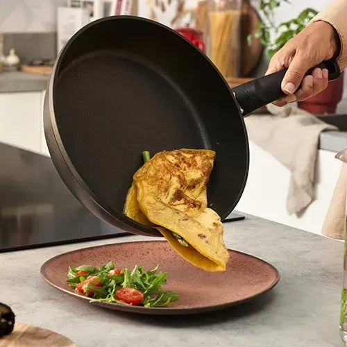 Eine Person serviert ein gefaltetes Omelett aus einer Fissler Antihaft-Pfanne auf einem Teller mit Salat in einer eleganten, modernen Küchenumgebung.
