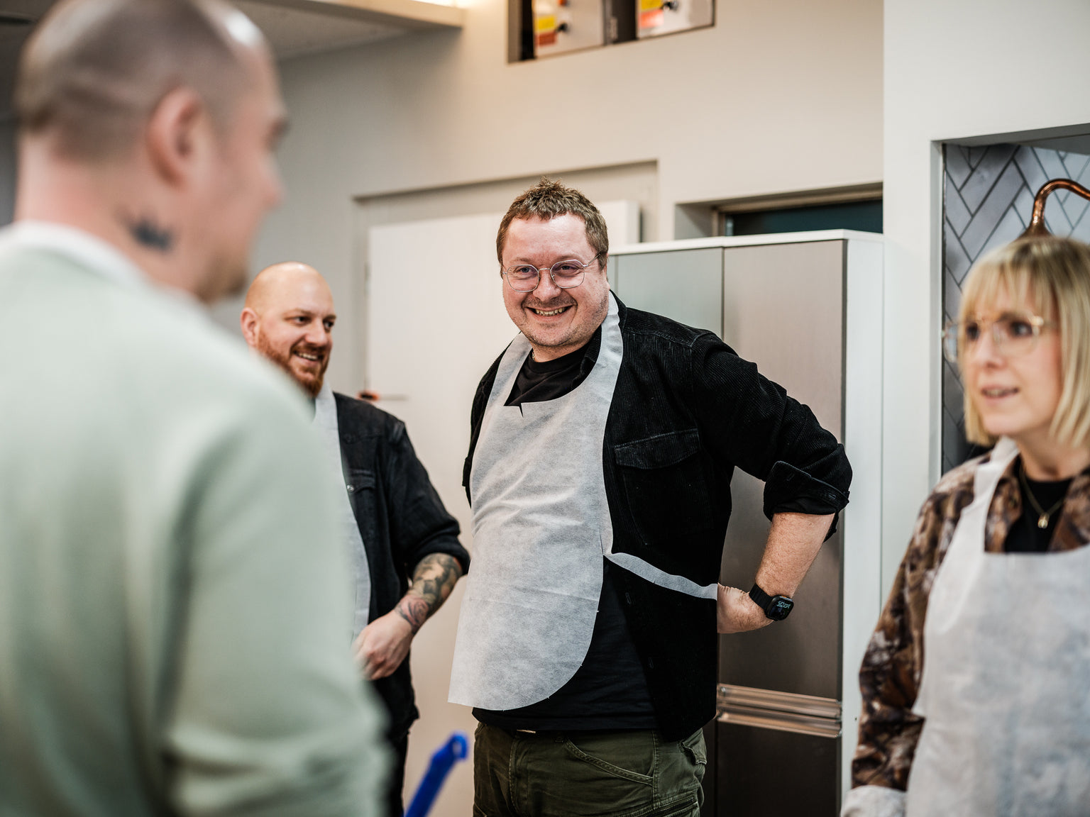 Drei Erwachsene sitzen in einer modernen Küche zusammen, lächeln und unterhalten sich. Der Mann in der Mitte, der eine Schürze und eine Brille trägt, steht neben glänzendem Fissler-Edelstahl-Kochgeschirr auf der Arbeitsplatte. Zu seiner Rechten hört eine Frau mit Brille aufmerksam zu, während links im Bild ein weiterer Mann teilweise zu sehen ist. Hochwertige Töpfe und Pfannen aus deutscher Fertigung verleihen ihrem gemeinsamen Kocherlebnis einen Hauch von zeitlosem Design und Langlebigkeit.