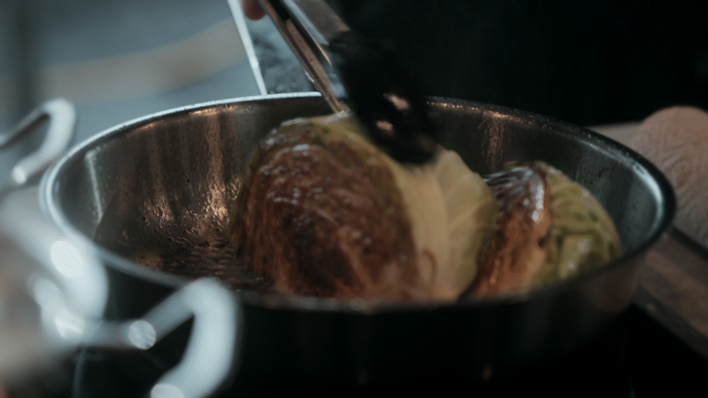 A person sears a halved cabbage, browning the cut sides, in a Fissler stainless steel pan using tongs. The pan’s high-quality German engineering ensures even heat distribution and perfect results.