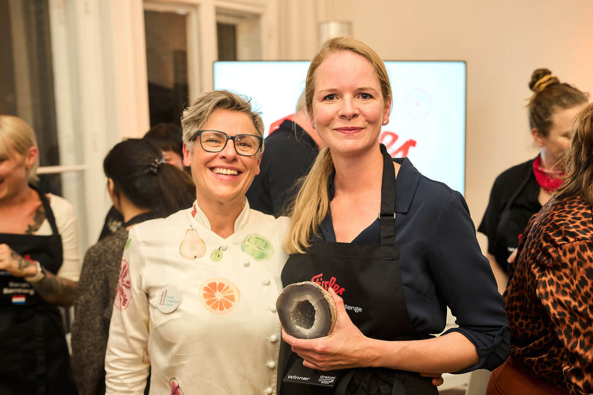 Two women smile at an event; one wears a Fissler apron and holds a high-quality German cookware lid, while guests mingle in the background.
