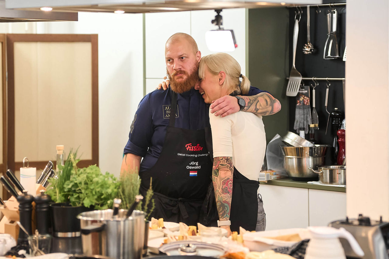 A chef in uniform hugs a distressed woman in a kitchen, surrounded by Fissler stainless steel pots and cookware on the counter.