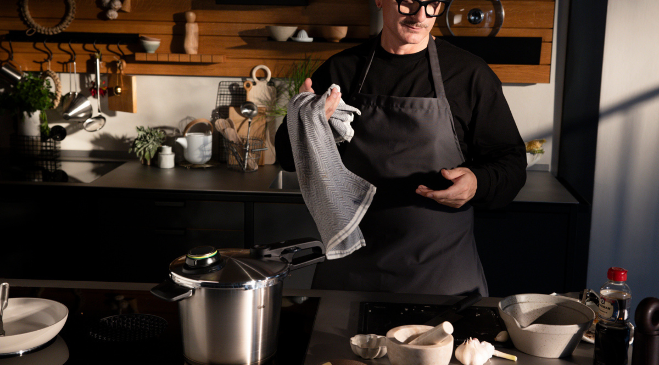 Eine Person in einer dunklen Küche steht neben einem Fissler Schnellkochtopf auf dem Herd, umgeben von hochwertigem Kochgeschirr und frischen Zutaten.