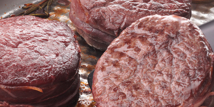 Three thick pieces of raw red meat begin to fry in a high-quality Fissler stainless steel pan, juice glistening on the surface.