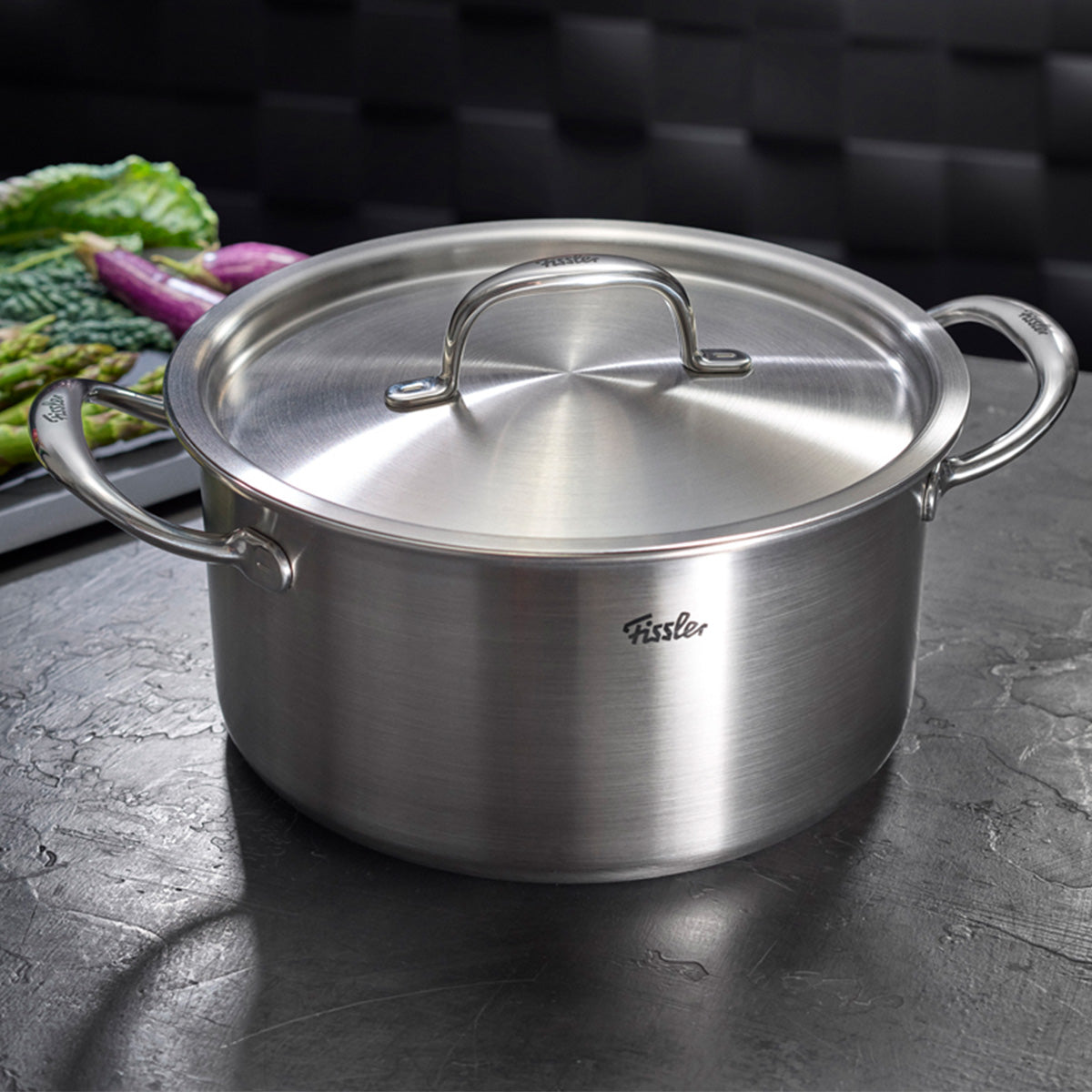 Das Fissler M5 Pro-Ply® 4-teilige Set mit Metalldeckeln, gefertigt aus recyceltem Edelstahl “Made in Germany”, präsentiert sich auf einer dunklen Arbeitsplatte. Umgeben von stilvoll arrangiertem grünem und violettem Gemüse unterstreicht das innovative, präzise verarbeitete Kochgeschirr unsere Tradition für langlebige Qualität und zeitloses Design. Das vielseitige Set ist die ideale Ergänzung zu jedem Pfannenset und optimal für alle induktionsgeeigneten Küchen geeignet.