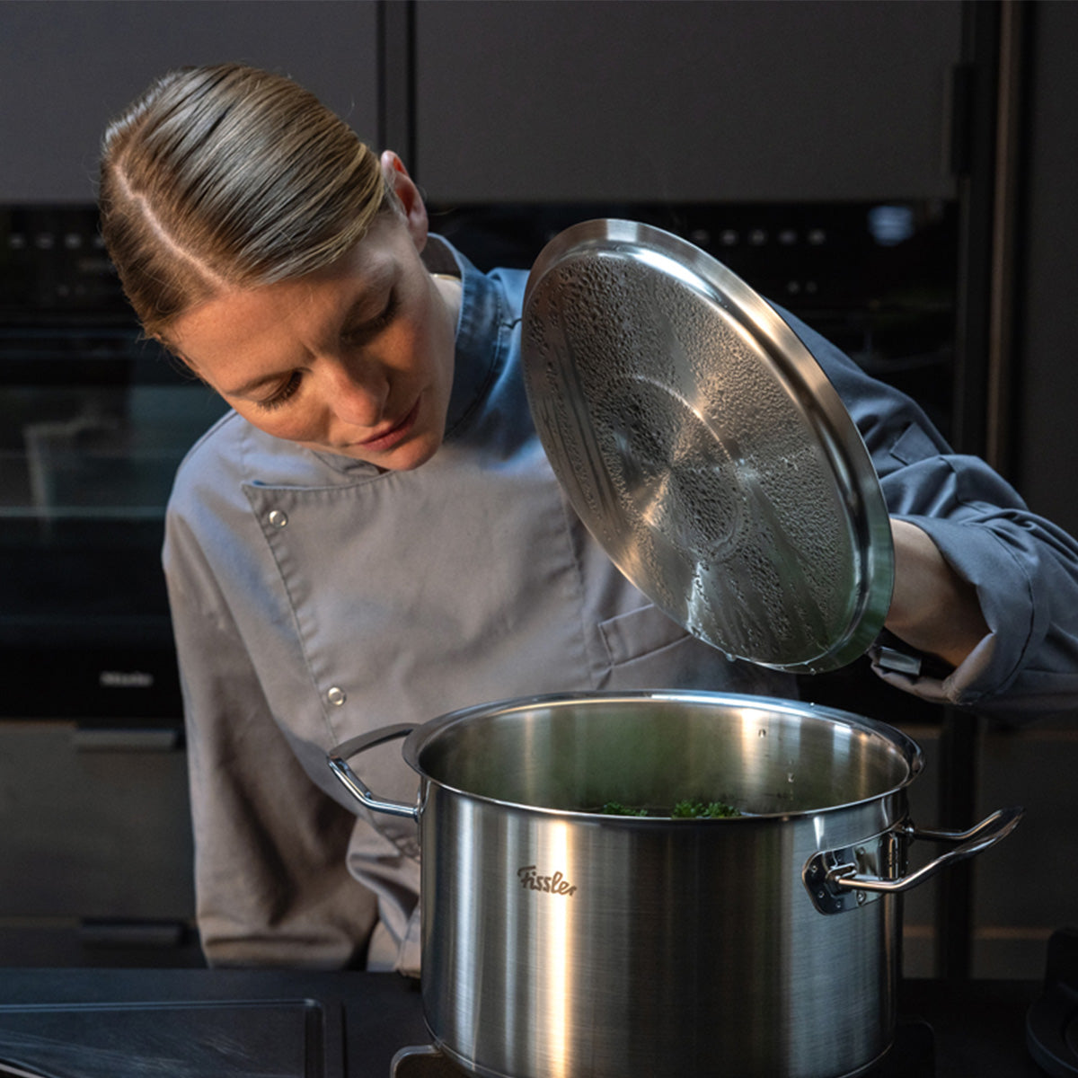 A chef in a modern kitchen lifts the lid of a stainless steel pot from the Original Profi Collection® by Fissler, known for German precision.