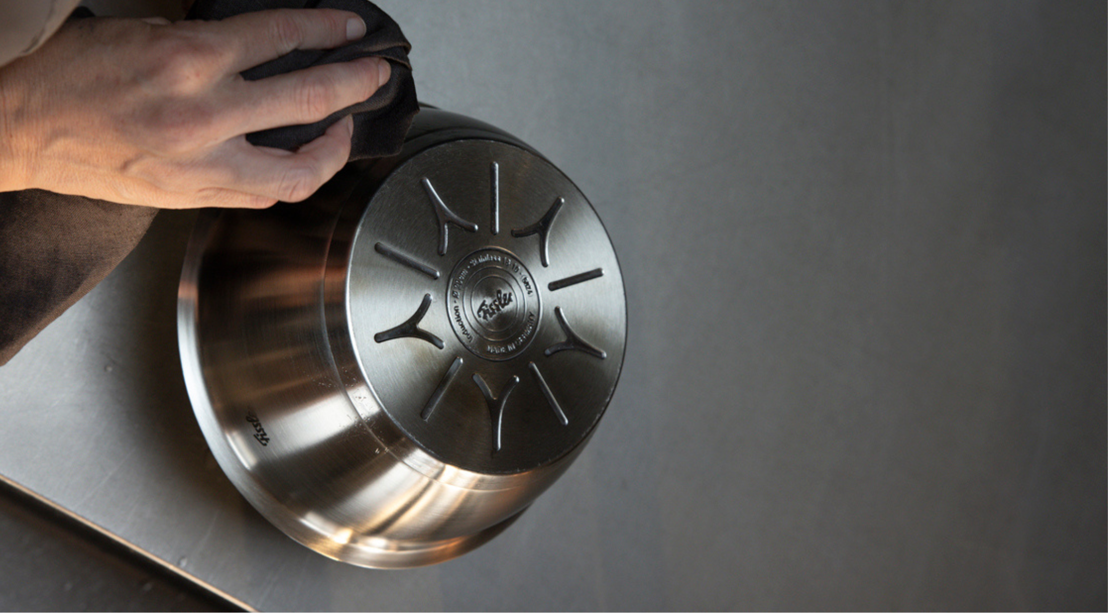 A hand uses a cloth to polish the base of a Fissler stainless steel bowl on a gray surface. The bowl features Fissler’s signature circular pattern and engraved logo, reflecting superior German engineering and timeless design.