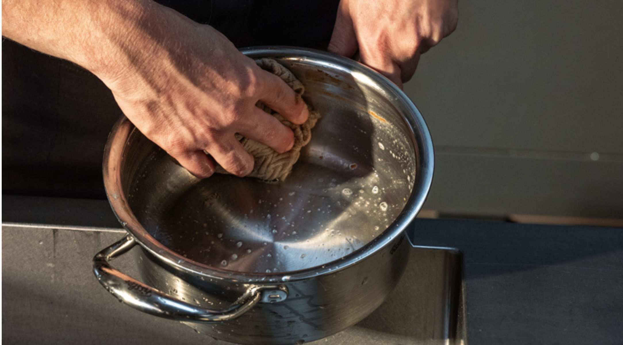 A person carefully cleans a Fissler stainless steel pot with a sponge and soapy water over a sink, showcasing the durability and enduring quality of precision-engineered German cookware.