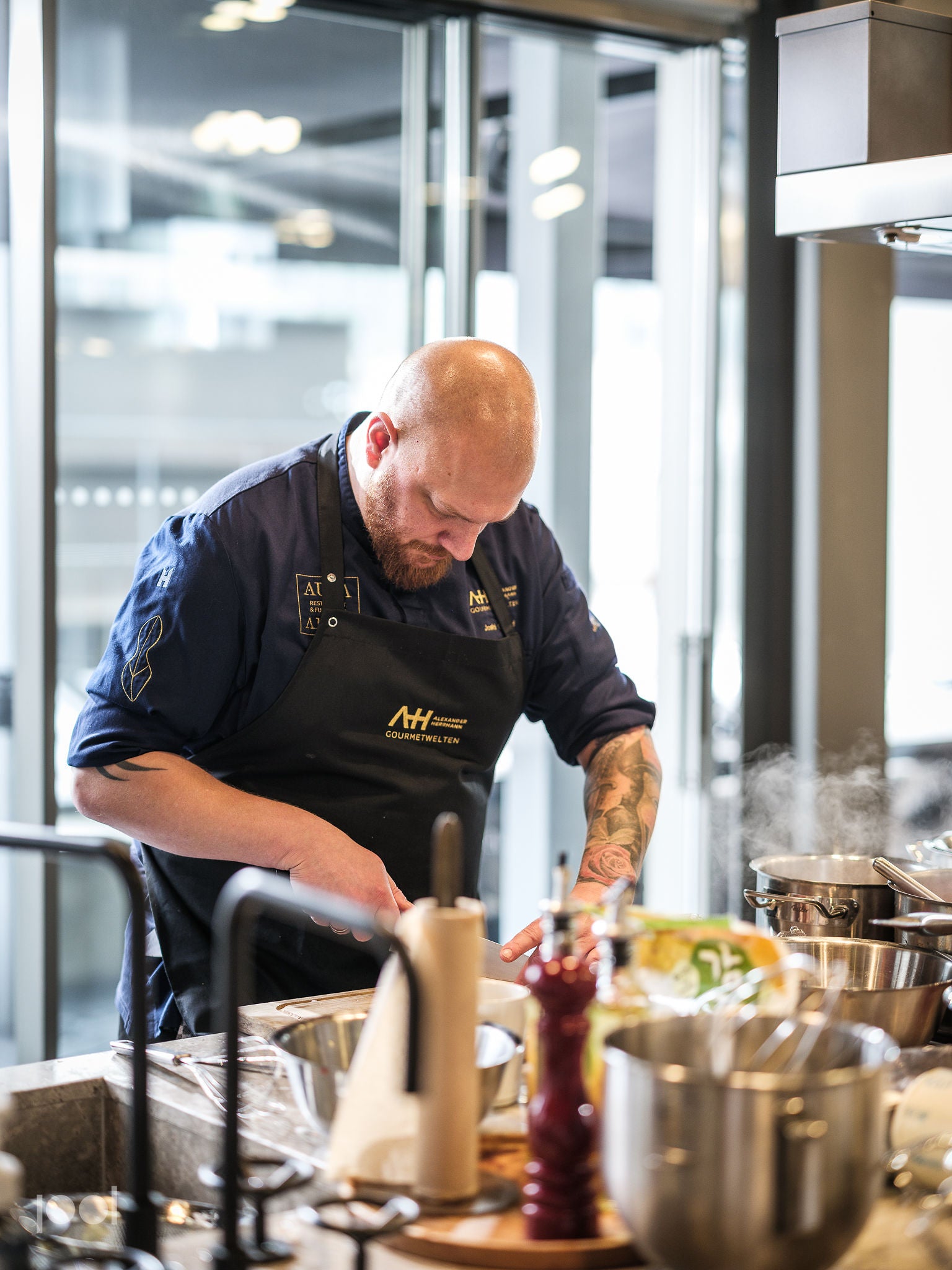 A chef in a blue apron prepares fresh ingredients at a kitchen counter, surrounded by premium Fissler stainless steel pots, pans, and utensils. The contemporary, sunlit kitchen showcases the precision engineering and timeless design of Fissler’s German-made cookware, with steam rising gently from durable pots as the chef cooks.