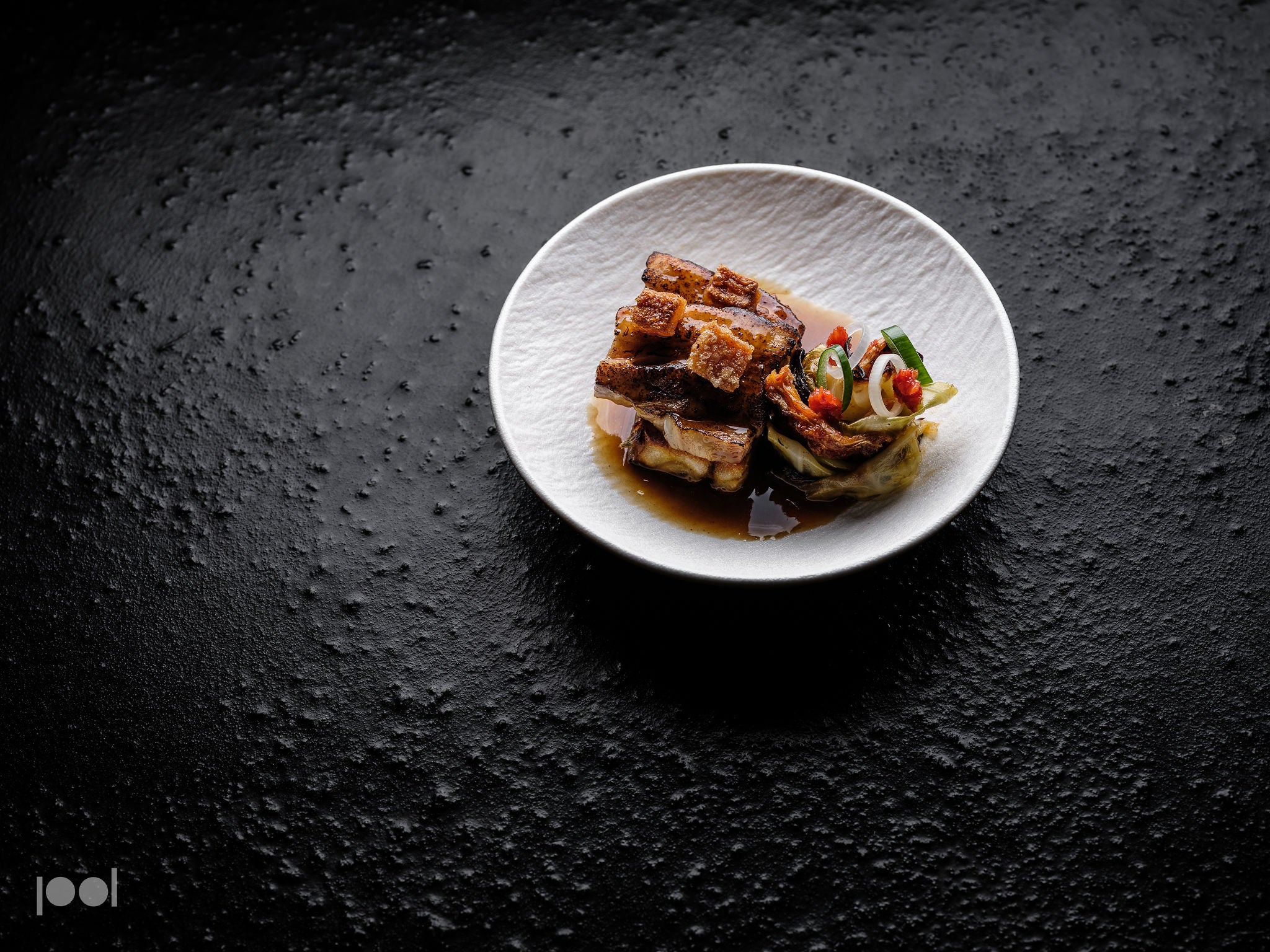 A white plate showcasing braised pork belly and vibrant vegetables is artfully presented atop a textured black surface, illuminated by dramatic lighting that accentuates every detail—capturing the culinary excellence achievable with Fissler’s precision-engineered, high-quality cookware.