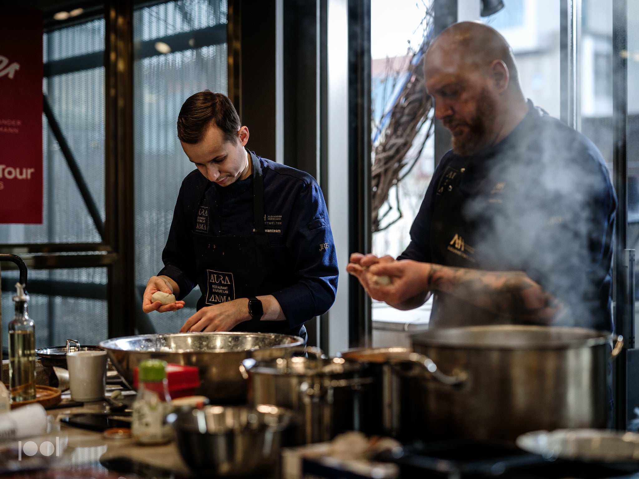 In a bright, naturally lit professional kitchen, two chefs in dark uniforms prepare food using premium Fissler stainless steel cookware. One chef expertly shapes dough beside gleaming Fissler pots and pans, while the other tends to steaming pressure cookers on the stove. The counter showcases a selection of durable Fissler kitchen utensils and fresh ingredients, reflecting precision engineering and timeless German design.