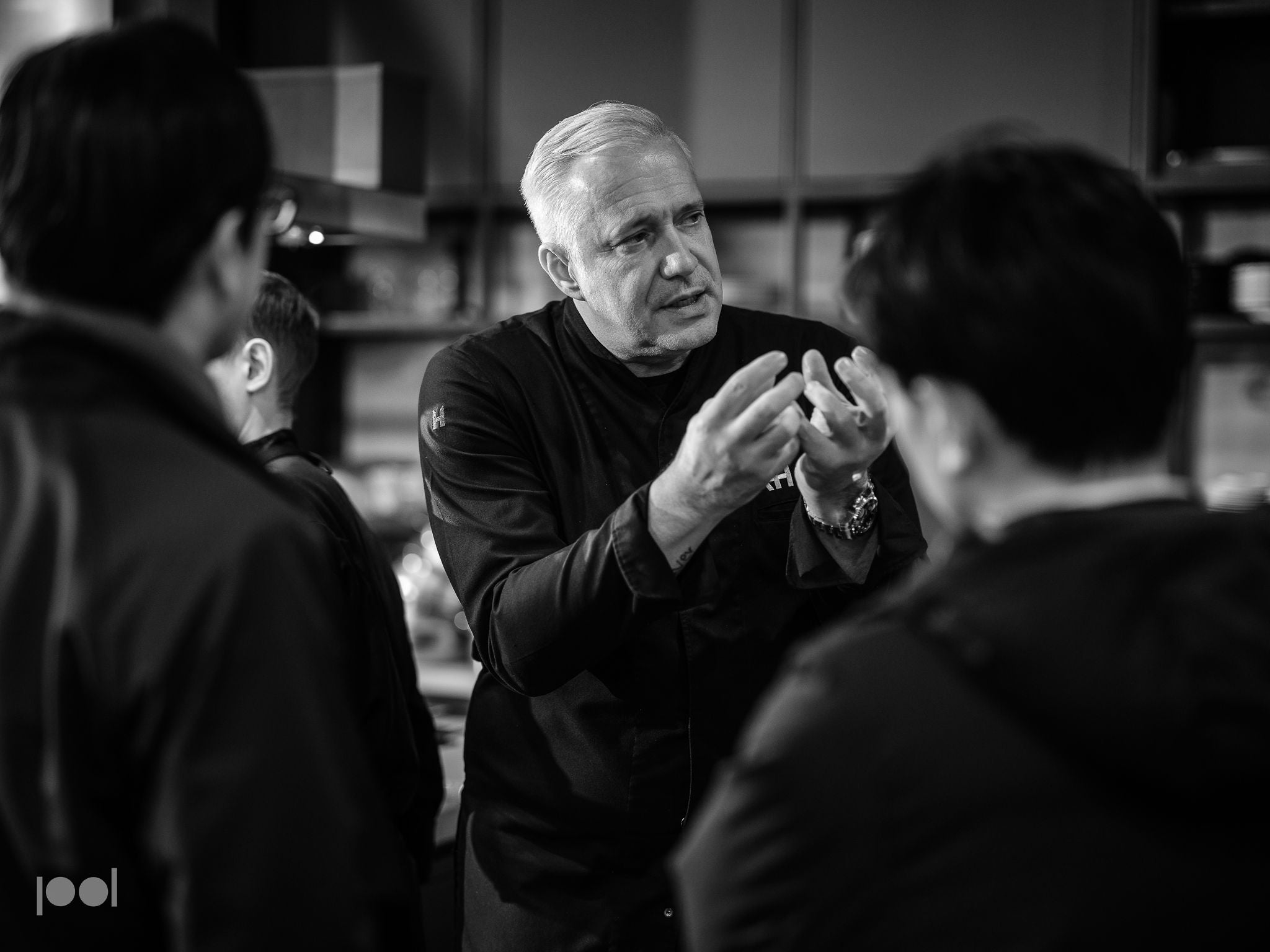 A chef in a dark uniform gestures with his hands while engaging two people in a kitchen, highlighting precision and expertise. The black-and-white scene centers on the chef’s expressive explanation, with sleek Fissler stainless steel pots and cookware elegantly arranged, reflecting the brand’s heritage of German engineering, durability, and timeless design.