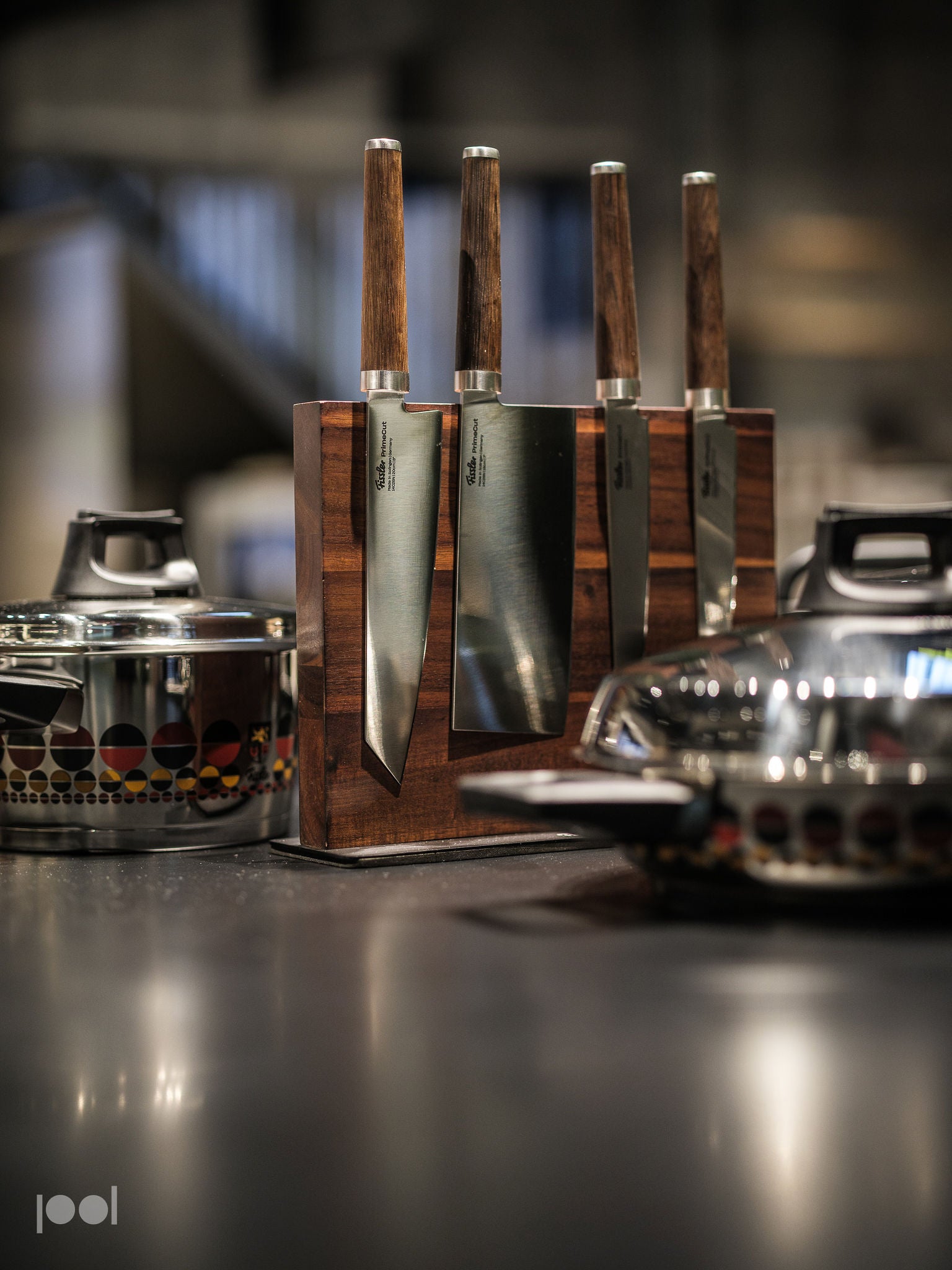 A set of four precision-engineered kitchen knives with elegant wooden handles rests in a wooden knife block on the countertop, surrounded by premium Fissler stainless steel cookware featuring timeless patterned designs—a showcase of German craftsmanship and innovation.