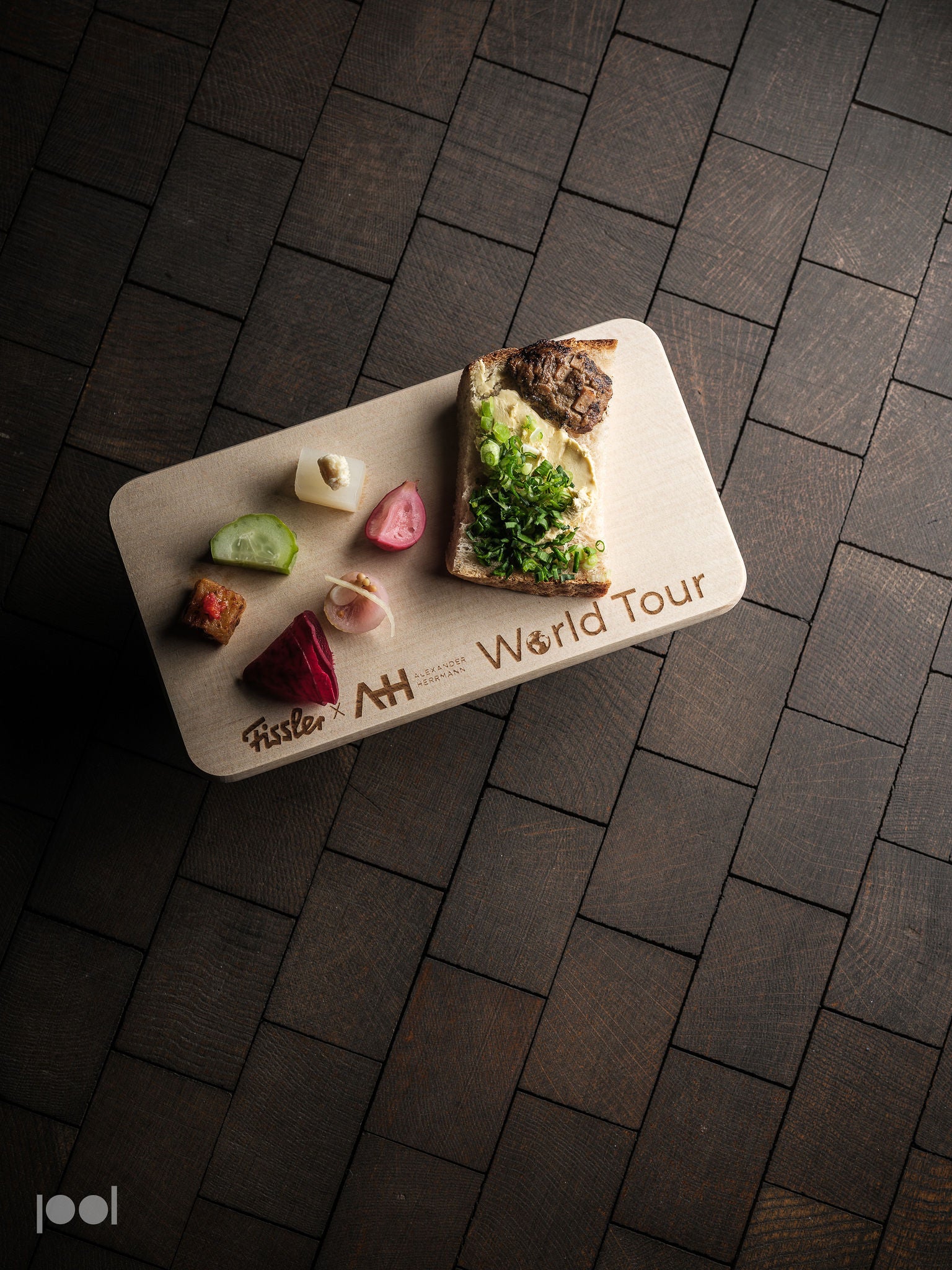 On a dark tiled surface, a precision-crafted wooden board engraved with "World Tour" showcases a slice of bread topped with fresh green herbs and six small, artfully arranged colorful food samples—an elegant presentation that reflects Fissler’s commitment to quality, durability, and timeless design in kitchen essentials.