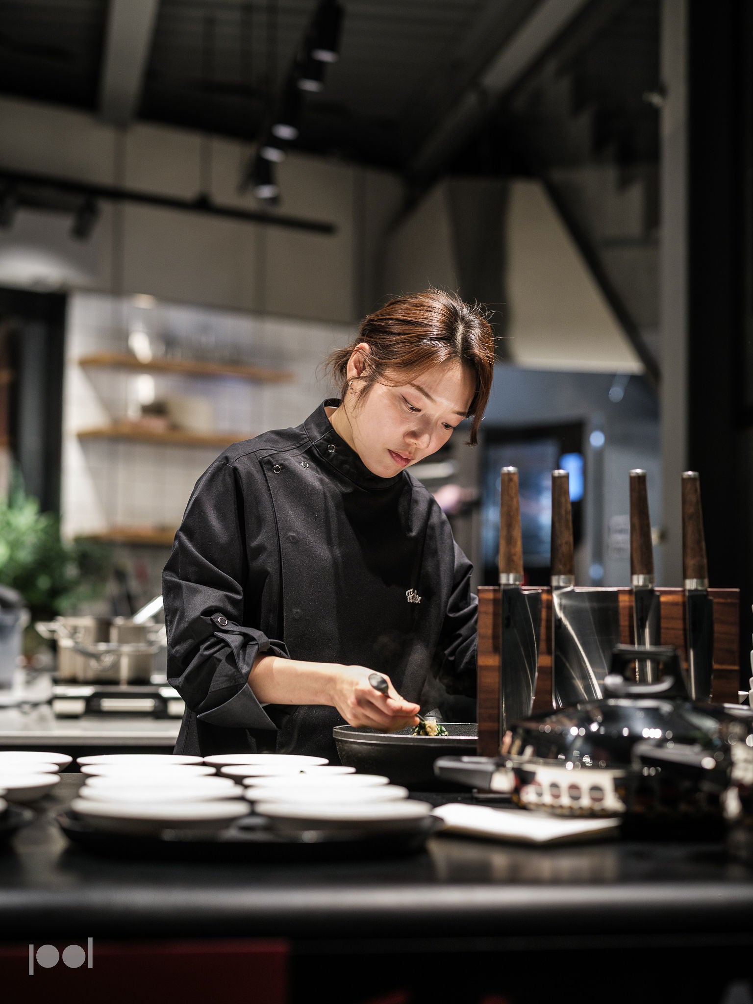 A chef in a black uniform prepares food with precision in a modern kitchen, focusing intently. Stainless steel Fissler pots, pans, and expertly crafted knives are arranged on the counter alongside other high-quality Fissler kitchen tools, reflecting German engineering and timeless design.